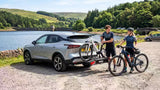 Two people unloading ebikes from a Thule car bike rack, in a car park beside a scenic reservoir.