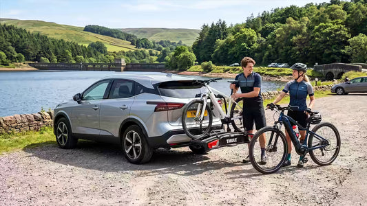 Two people unloading ebikes from a Thule car bike rack, in a car park beside a scenic reservoir.
