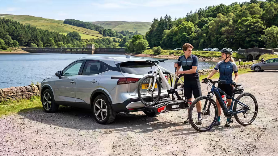 Two people unloading ebikes from a Thule car bike rack, in a car park beside a scenic reservoir.