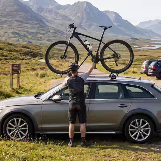 A solo mountain biker unloading his bike from a roof-mounted cycle carrier rack on an Audi estate car in a remove car parking spot in the Scottish Highlands