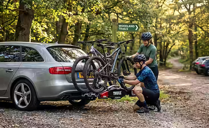 Two people loading bicycles onto a car in a wooded area with a 'Woodland Trails' sign.