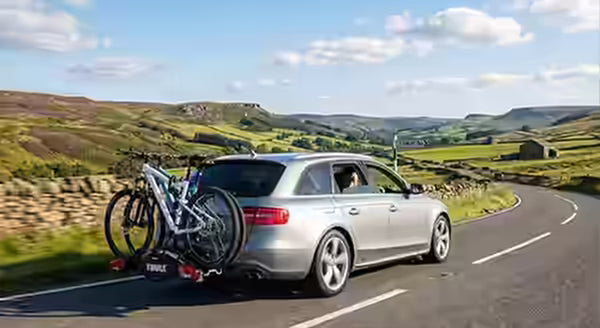 Silver Audi estate car with a towbar mounted bike carrier and two bikes, driving along a peak district road