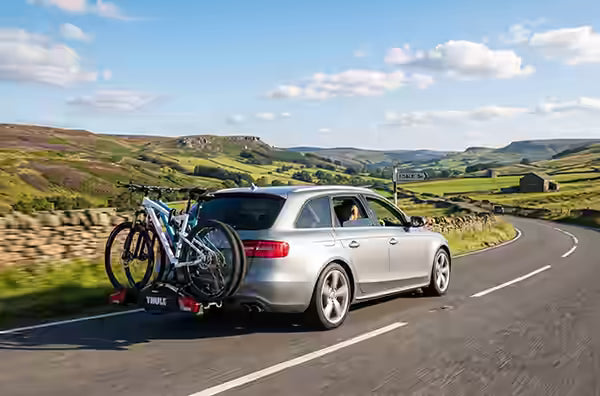 Silver Audi estate car with a towbar mounted bike carrier and two bikes, driving along a peak district road
