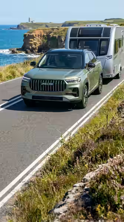 Green SUV towing a caravan on a scenic coastal road.