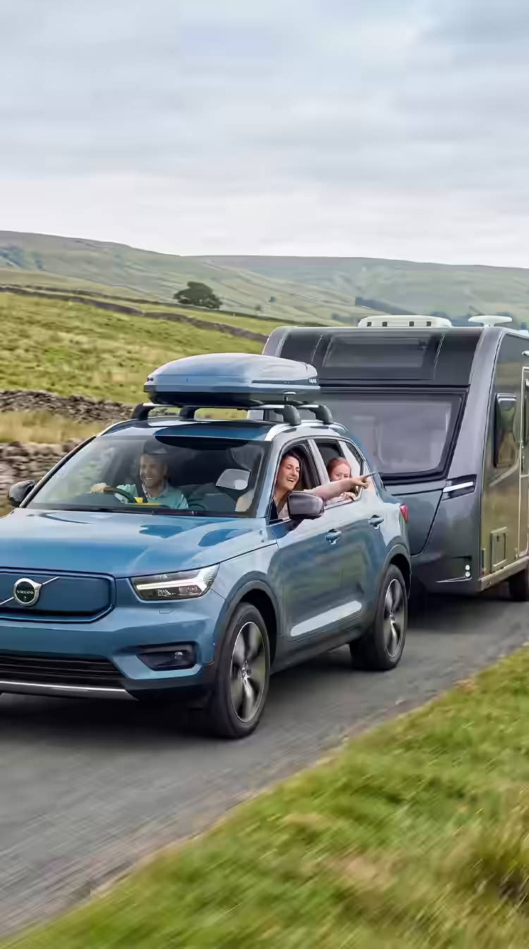 A happy family heading off on a caravanning holiday in the UK in a Blue Volvo XC40 SUV towing a sleek modern caravan along a winding country road