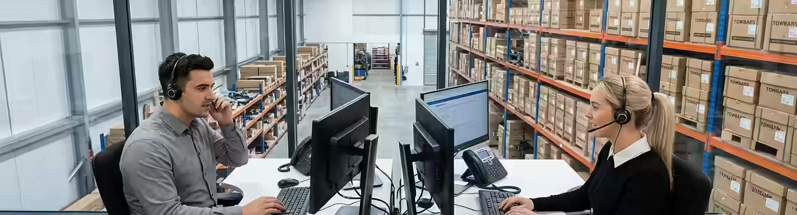 Two colleagues in an office overlooking a towbar warehouse, one at a computer desk and the other on a phone call.